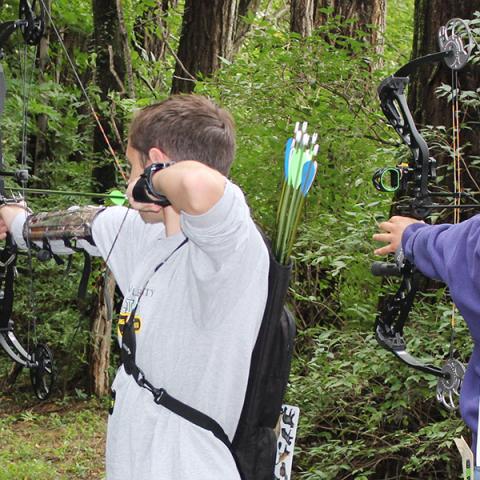 2 teens shooting bow and arrows in woods